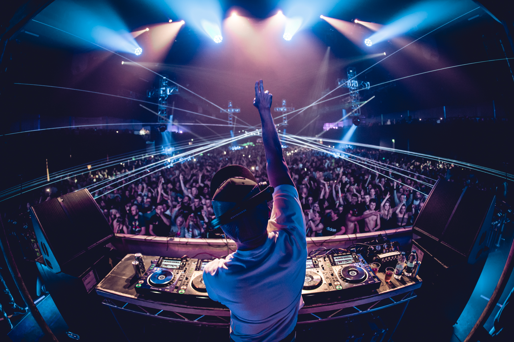 Image of a man from behind on DJ decks with his arm up to a full room of people. the image is mostly purple with lasers 